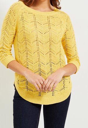 Femme portant un pull tricoté jaune avec un motif en dentelle et des manches trois-quarts, associé à un jean bleu foncé, les mains croisées devant.