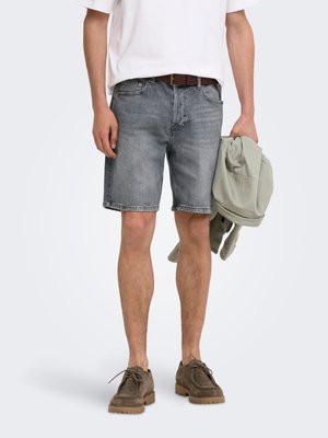 Homme portant un short en denim gris, une chemise blanche, une ceinture marron et des chaussures marron, tenant une veste vert clair dans la main droite.