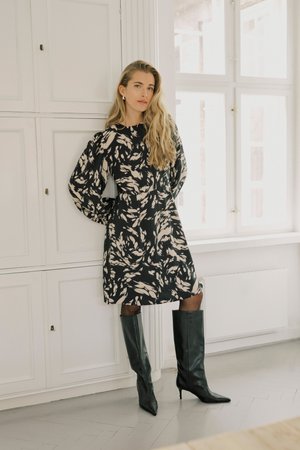 Frau mit langen blonden Haaren, die ein schwarz-beiges gemustertes Kleid und kniehohe schwarze Stiefel trägt, steht drinnen in der Nähe von weißen Schränken und einem Fenster.