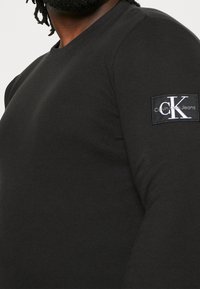 T-shirt noir à manches longues en tissu texturé, avec un petit patch logo CK sur la manche. Design simple et ajusté.