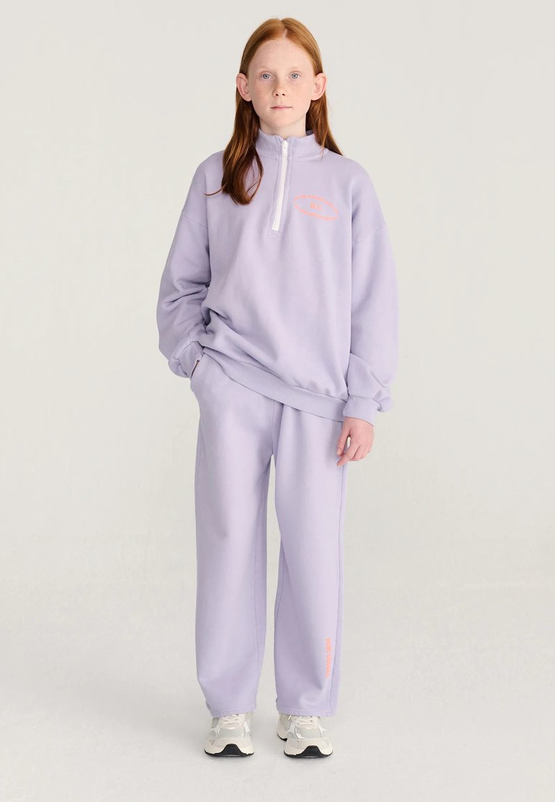 Jeune personne aux longs cheveux roux portant un sweat-shirt violet clair et un pantalon assorti avec des baskets blanches, debout devant un fond uni.