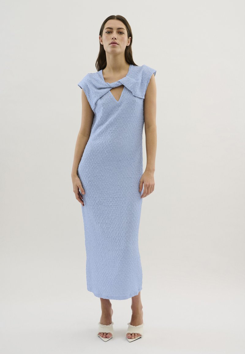 Rochie maxi texturată, de culoare albastru deschis, cu decolteu în V și detalii în formă de fundă; siluetă ajustată cu mâneci scurte, purtată cu pantofi cu vârf descoperit.