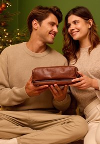 Pár sedící u vyzdobeného vánočního stromečku si vyměňuje hnědou koženou tašku, usmívají se na sebe a mají v očích vřelost.