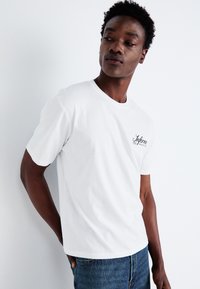 Witte katoenen t-shirt met een ronde hals en korte mouwen, voorzien van een zwart scriptlogo op de linkerborst.