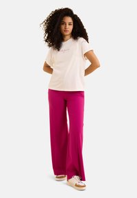Camiseta crema con mangas enrolladas que presenta texto bordado en rosa, combinada con pantalones de pierna ancha de color rosa brillante y zapatillas beige con detalles en blanco.