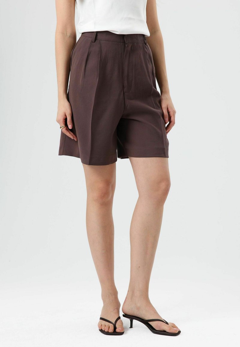 Bruine shorts met hoge taille, plooien, een getailleerde pasvorm en een enkelhoge lengte, gecombineerd met zwarte sandalen met bandjes. Gladde stoftextuur.