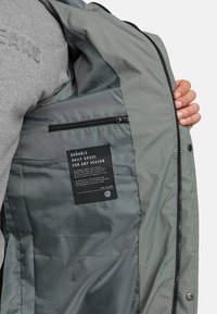 Grijze jasinterieur met gladde stof, voorzien van een met rits gesloten zak en een label met instructies. Zwarte rits en drukknopdetails zichtbaar.