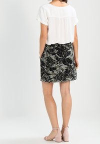 Chemisier blanc à manches courtes associé à une jupe mi-longue arborant un motif floral noir sur fond vert ; porté avec des sandales couleur vieux rose.