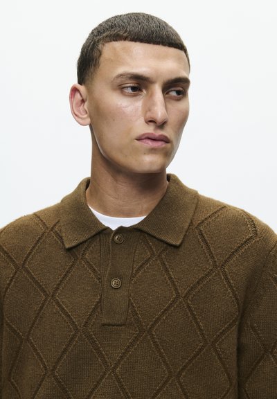Polo en maille marron avec un motif en losanges, doté d'une patte de boutonnage à trois boutons et d'un col court. Texture douce, porté par-dessus un t-shirt blanc.