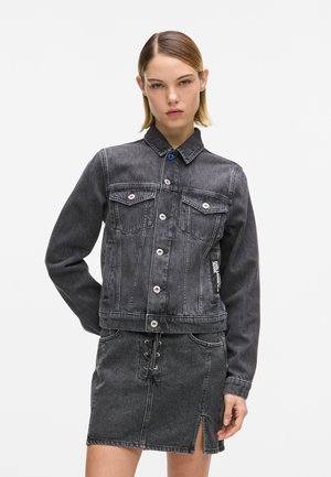 Jonge vrouw met kort haar, gekleed in een zwarte spijkerjasje en bijpassende rok met een split aan de voorkant, tegen een effen achtergrond.