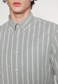 Koszula zapinana na guziki z jasnoszarego materiału w białe pionowe paski. Posiada kołnierzyk, cztery guziki oraz kieszeń na piersi.