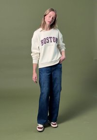 Femme portant un sweat-shirt blanc "Boston", un jean large bleu et des baskets blanches, debout devant un fond vert uni.