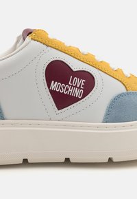 Fehér sneaker sárga és kék velúrbetétekkel. Bordó szív logóval rendelkezik, amelyen az áll: "LOVE MOSCHINO." Mintás talp a tapadásért.
