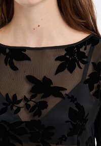 Woman’s neck and shoulders wearing a black sheer top with velvet floral patterns and a black undergarment visible beneath.