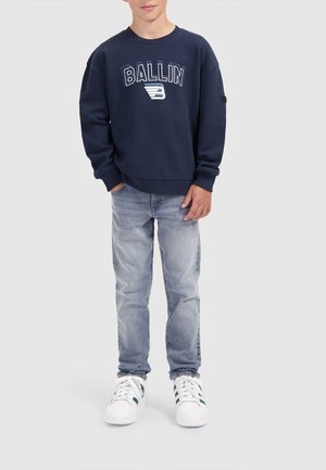 Garçon portant un sweat-shirt bleu marine avec le texte "BALLIN", un jean délavé clair et des baskets blanches, debout devant un fond uni.