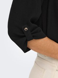 Chemise noire à manches trois-quarts avec détail de patte boutonnée portée avec un pantalon beige clair sur le bras et la taille d'une personne.