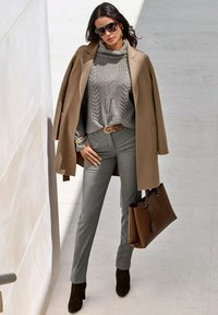 Camelkleurige jas, grijze kabelgebreide trui, grijze slimfit broek, bruine leren handtas en bruine enkelschoenen. Grote zwarte zonnebril.