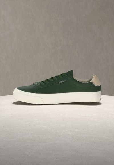 Σκούρο πράσινο παπούτσι τύπου sneaker χαμηλού ύψους με λευκή σόλα και μπεζ ταμπελάκι στη φτέρνα, τοποθετημένο σε ανοιχτή επιφάνεια με υφή και φόντο απλό γκρι.