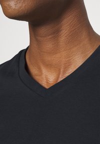Close-up af nakke og øvre bryst iført sort V-hals t-shirt mod ensfarvet lyst baggrund.