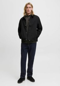 Giacca leggera nera con colletto, bottoni a pressione e design semplice, indossata sopra una maglietta scura e abbinata a jeans e scarpe scure.