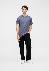 Μωβ βαμβακερό μπλουζάκι με στρογγυλή λαιμόκοψη και κοντά μανίκια, με μικρό κίτρινο λογότυπο. Συνδυασμένο με μαύρο παντελόνι και λευκά αθλητικά παπούτσια.