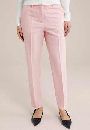 Nainen pukeutuneena vaaleanpunaiseen suoran lahkeen housuun, jossa on etuvakotus, yhdistettynä mustiin kärjestä teräväkärkisiin kenkiin ja valkoiseen pitkähihainen toppiin.