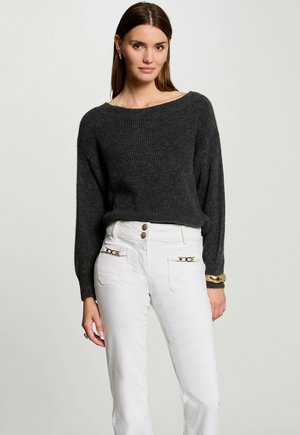 Maglione in grigio carbone con scollatura ampia e maniche lunghe, abbinato a pantaloni bianchi con tasche frontali accentuate da dettagli dorati.