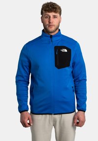 Veste softshell bleue avec une poche poitrine zippée noire, deux poches latérales zippées et des coutures contrastées. Texture lisse, design ajusté.