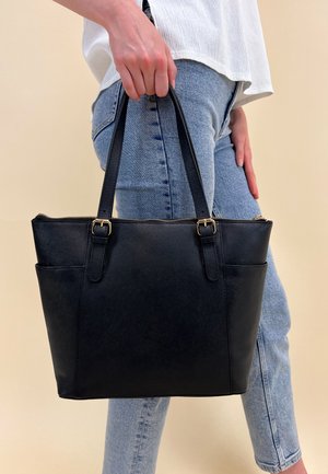 Black leather tote bag with gold hardware, zip closure, structured shape, and dual handles. Side pocket visible. Worn with denim jeans.