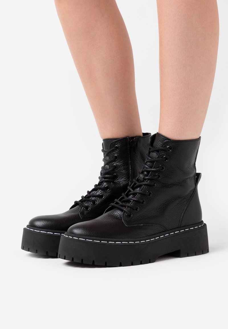 Bottes de combat en cuir noir à lacets avec semelles plates-formes épaisses, portées sur des jambes nues sur fond blanc.
