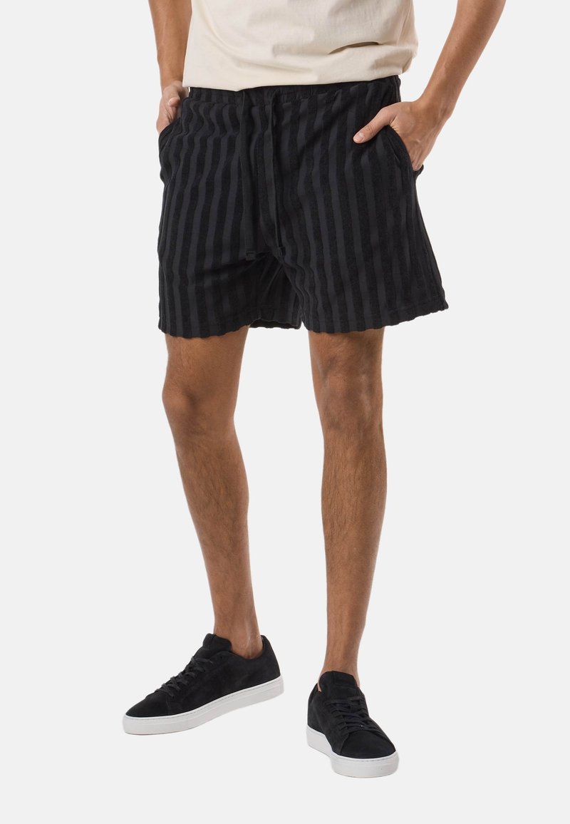 Homme portant un short noir à rayures, des baskets noires à semelles blanches et une chemise beige, debout les mains dans les poches.