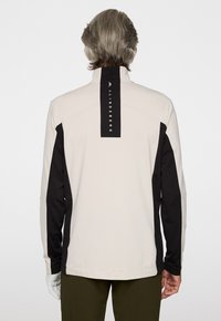 Maglia leggera grigia a maniche lunghe con accenti neri e colletto alto; presenta un'etichetta nera distintiva con testo bianco sul retro.