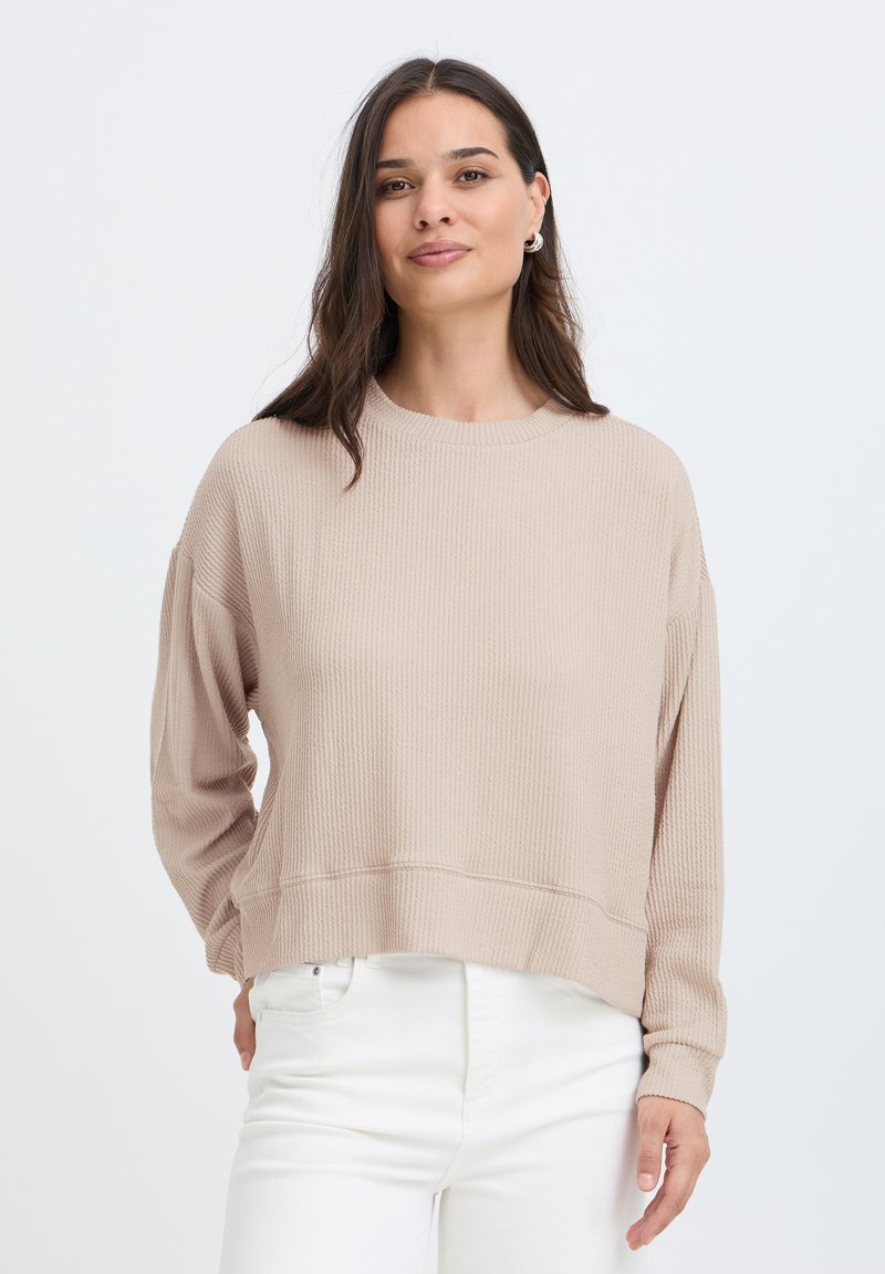 Femme aux longs cheveux foncés portant un pull côtelé beige et un pantalon blanc, debout devant un fond clair uni.