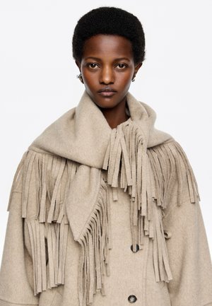 Manteau en laine beige avec des franges autour du col, présentant une finition texturée et de grands boutons, créant un look superposé.