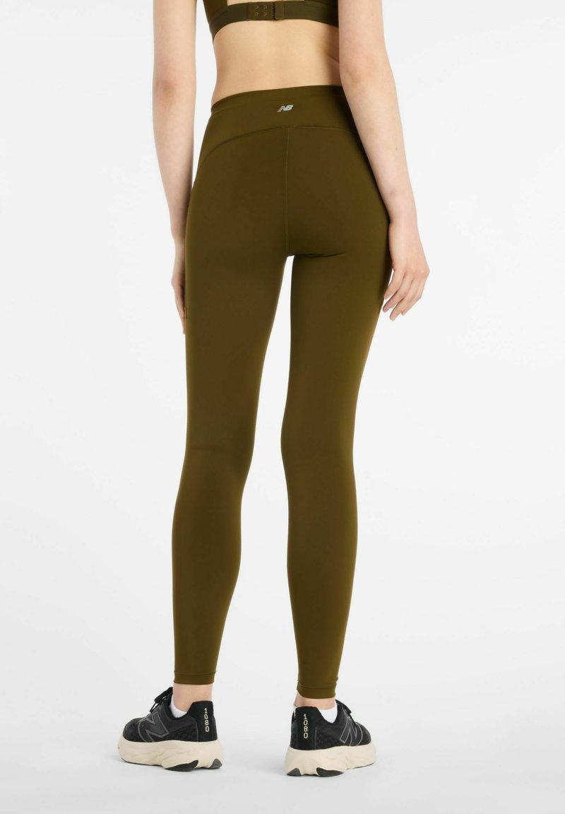 Olivgröna leggings med hög midja, stretchigt tyg och flatlock-sömmar. Bärs med svarta träningsskor med vita detaljer.