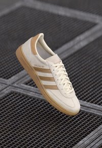 Sapatilhas bege com parte superior em lona texturizada, sola em borracha de goma e três riscas castanhas. Apresenta atacadores creme e a marca "SPEZIAL" na lateral.