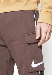 Bruna sweatpants med en sidocargo-ficka, vitt Nike-logotyp och svarta detaljer. Mjuk textur med en avslappnad passform.