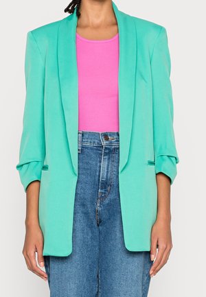 Personne portant un blazer turquoise sur un haut sans manches rose vif et un jean bleu taille haute devant un fond uni.