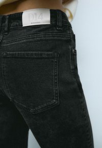 Crne traperice sa uskim krojem, s stražnjim džepom i etiketom označenom "D14" na pojasu. Meka tekstura i minimalni detalji šivanja.