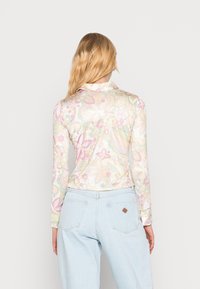 Mujer con cabello rubio largo vestida con una camisa de manga larga floral en tonos pastel y jeans de tiro alto azul claro, de espaldas contra un fondo liso.