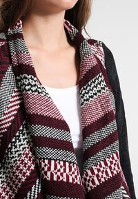 Stickad cardigan med ett röd, svart och vitt geometriskt mönster, med avslappnad passform och öppen framsida. Mjuk textur och lagerdesign.