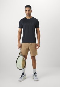 Fekete, rövidujjú póló, amelyen egy nagy, tónusos "BOSS" logó található, bézs rövidnadrággal és fehér térdzoknival párosítva, teniszütőt tartva.