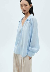 Blusa azzurro chiaro con scollo a V, maniche lunghe a sbuffo e polsini, realizzata in un tessuto morbido e fluido.
