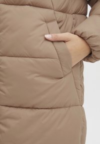 Brown puffer jacket with a smooth, shiny exterior and horizontal quilting. Features a side seam pocket with a hand partially inserted.
