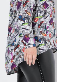 Tunika stampata multicolore che presenta icone della moda e testo su uno sfondo bianco, abbinata a leggings neri texturizzati con accenti di strass.