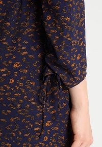 Granatowa bluzka z pomarańczowym wzorem zwierzęcym, z detalami w postaci bufkowatych rękawów i wiązaniami przy nadgarstku. Miękka, teksturowana tkanina.