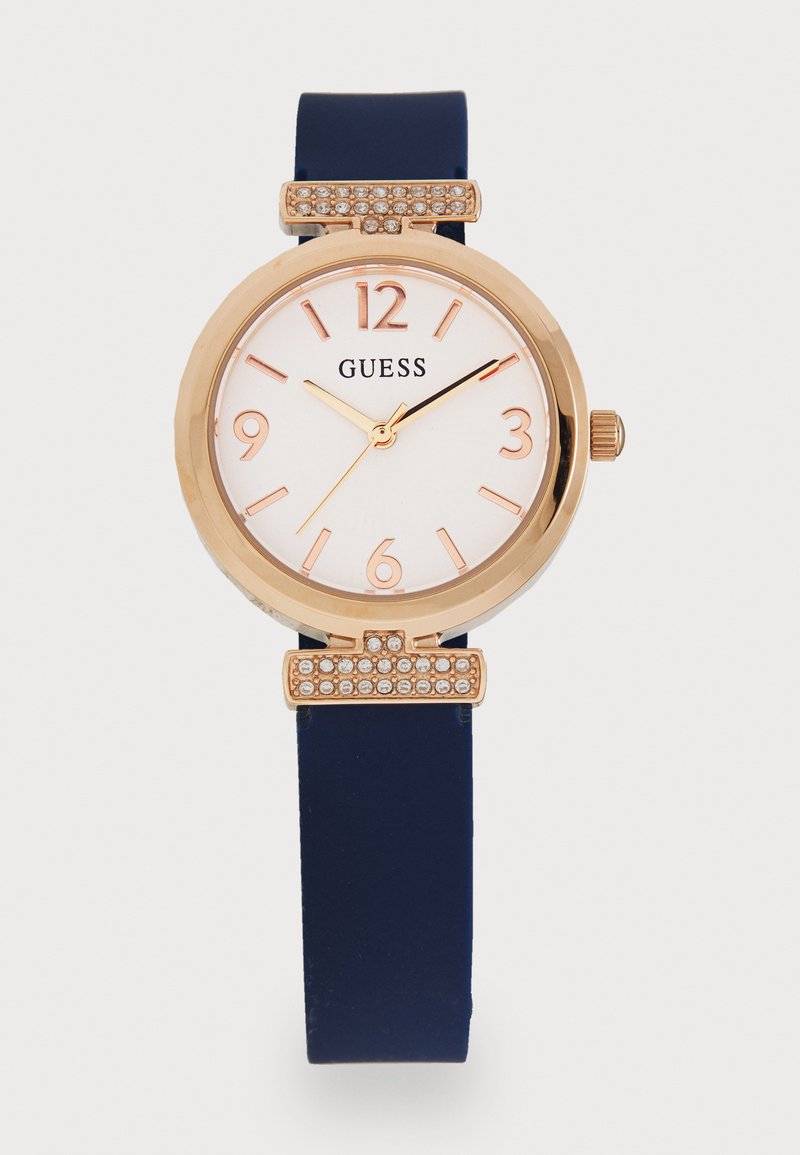 Gold-tone watch with a white dial, navy silicone strap, rose gold accents, and a crystal-embellished detail above the face.