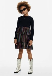 Haut noir à manches longues associé à une jupe à volants rayée arc-en-ciel et des bottes argentées montantes jusqu'aux genoux. Des lunettes de soleil noires complètent la tenue.