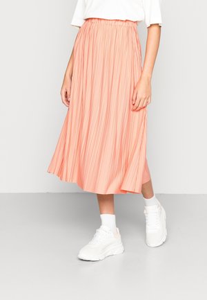 Femme portant une jupe midi plissée corail, un t-shirt blanc, des chaussettes blanches et des baskets blanches épaisses, debout sur un sol gris.