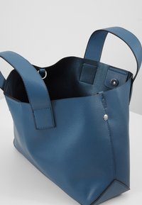 Modrá kožená taška přes rameno s texturovaným povrchem. Obsahuje dva pevné úchyty a bezpečné zapínání na patent. Interiér je hladký a prostorný.